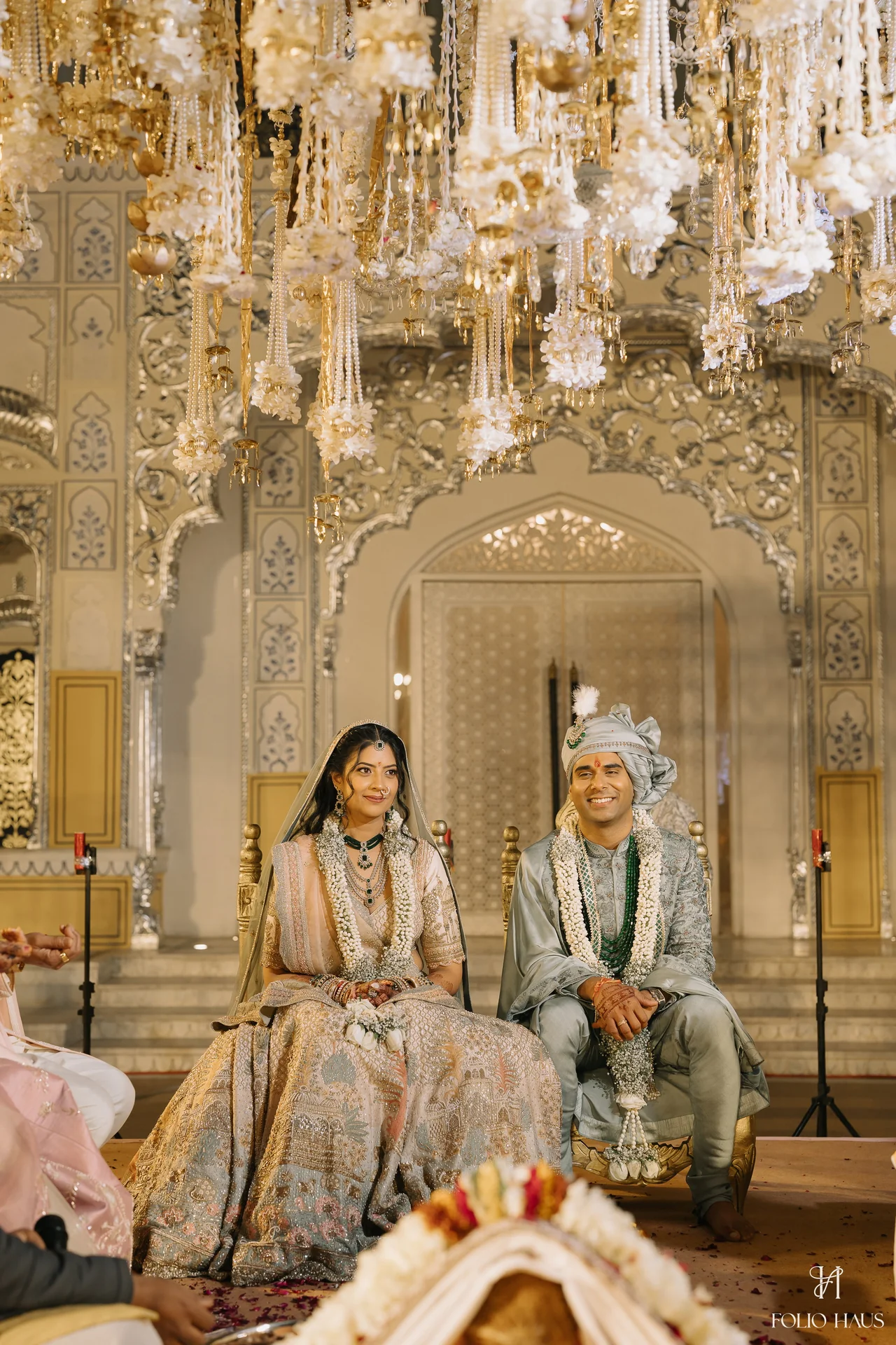 Jigna and Anurag seated together beneath hanging flowers during their ceremony.
